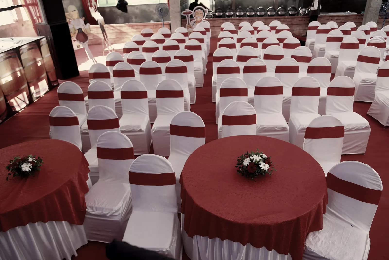 Rows of chair covers with crimson sashes behind two dressed round tables with small floral centres.