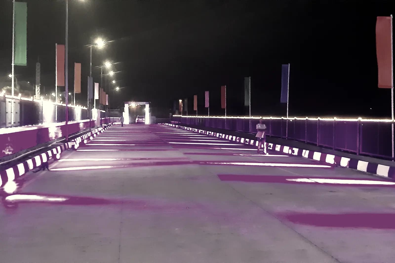 A wide approach road lit in deep violet at night, lined with banner flags and low ground lighting.
