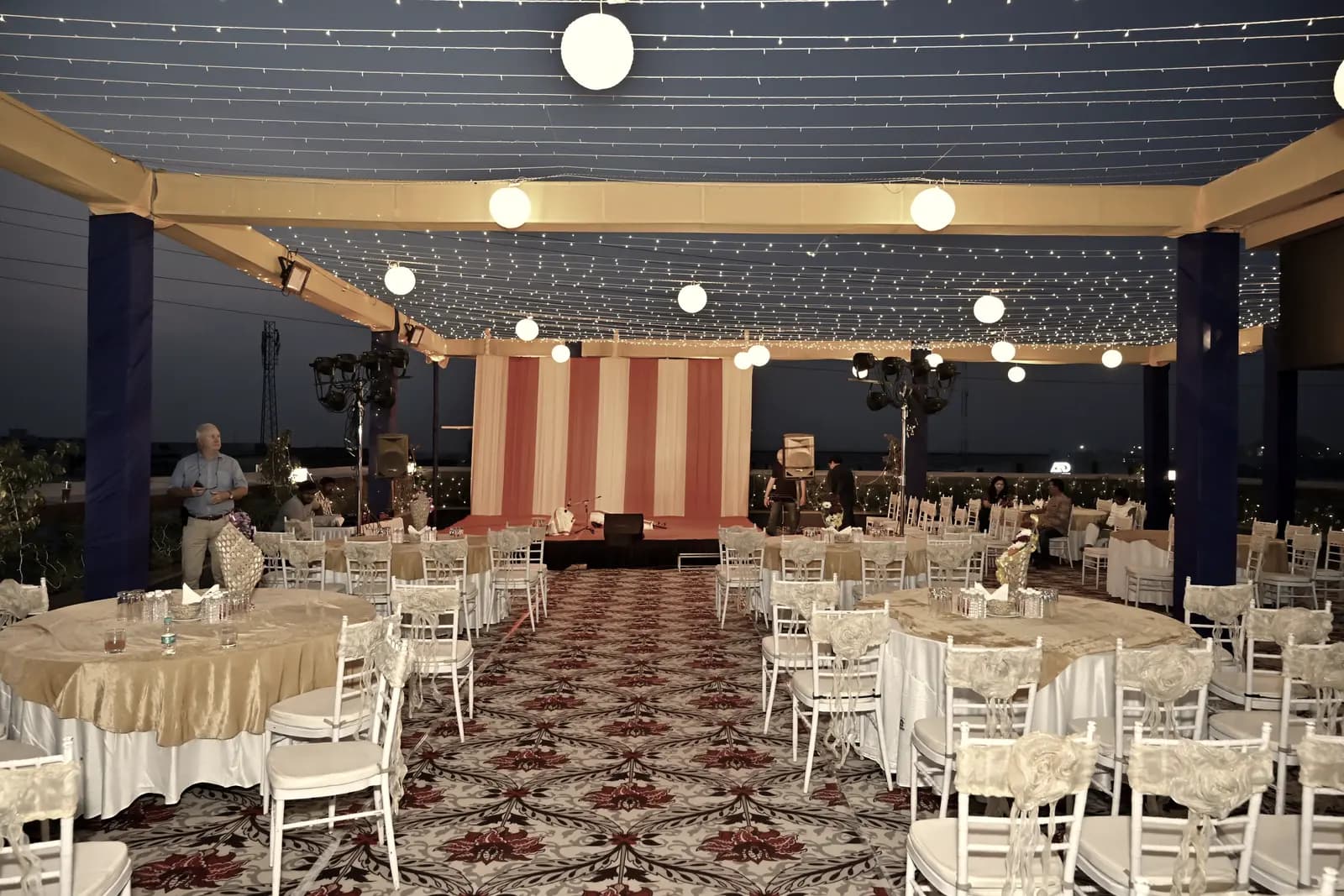 A rooftop dinner floor at dusk under canopy lights and fairy-light strands, tables laid and waiting.
